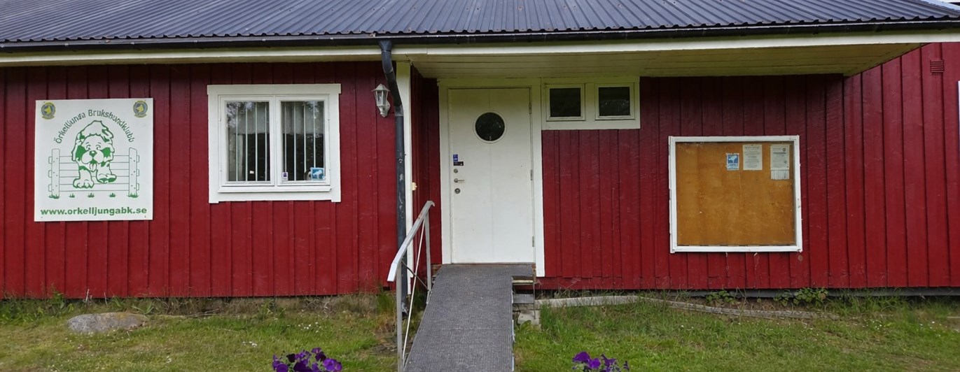 Röd klubbstuga med vita knutar och vit dörr. Blommor är placerade framför den handikappanpassade entrén.