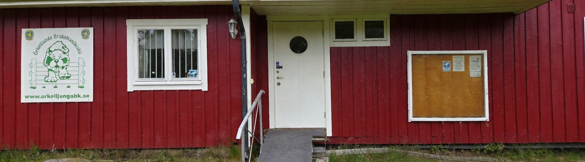 Röd klubbstuga med vita knutar och vit dörr. Blommor är placerade framför den handikappanpassade entrén.
