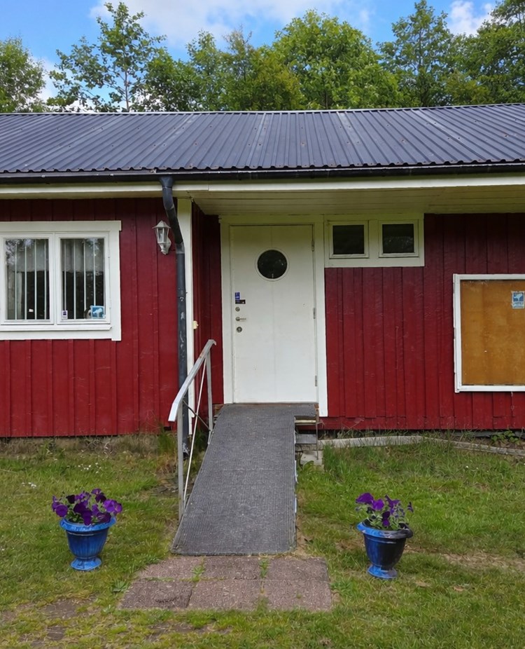 Röd klubbstuga med vita knutar och vit dörr. Blommor är placerade framför den handikappanpassade entrén.