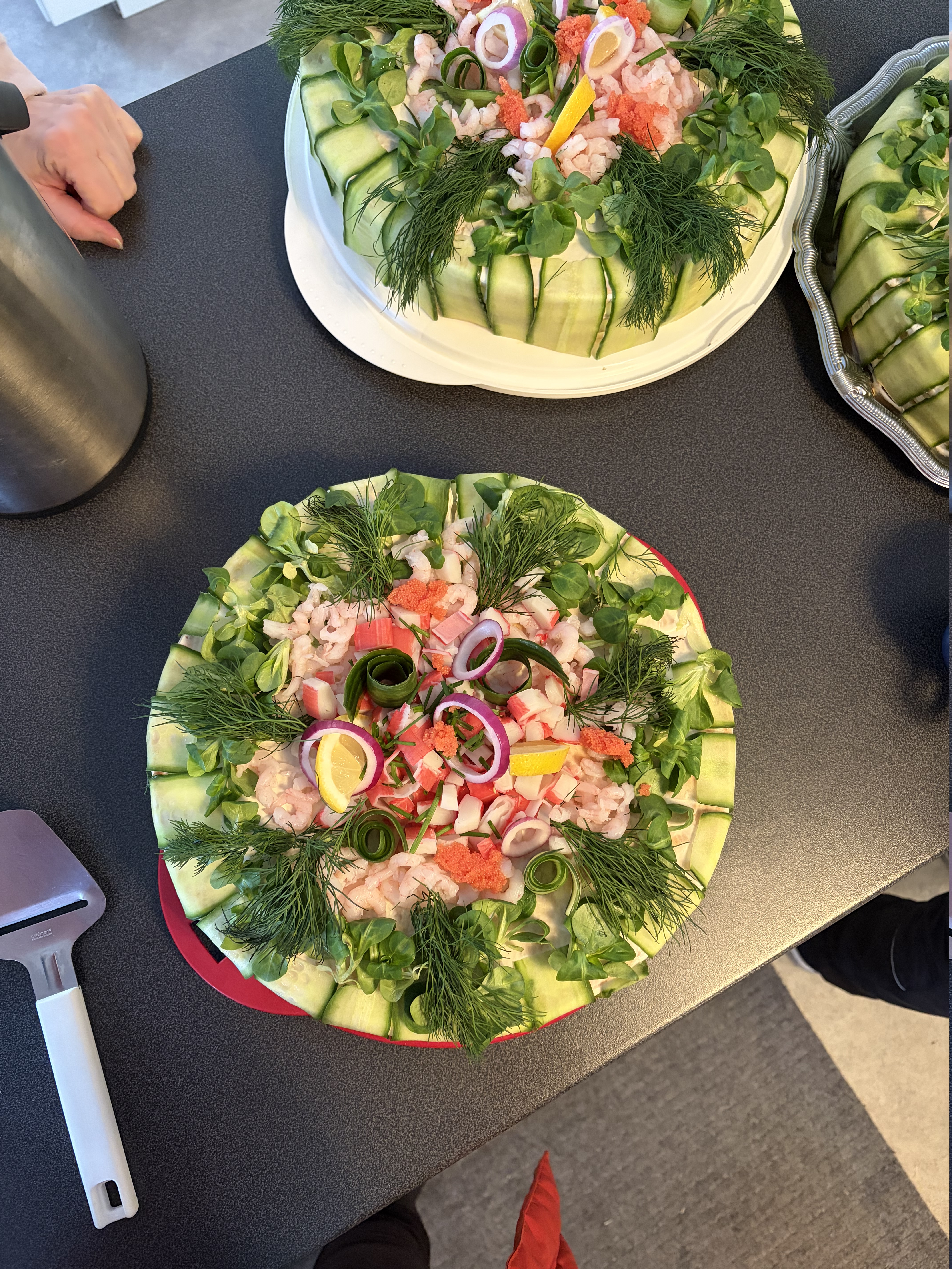En bild ovanifrån på två runda smötgåstårtor garnerade med gurka, räkor, dill, citron, stenbitsrom och purjolök.