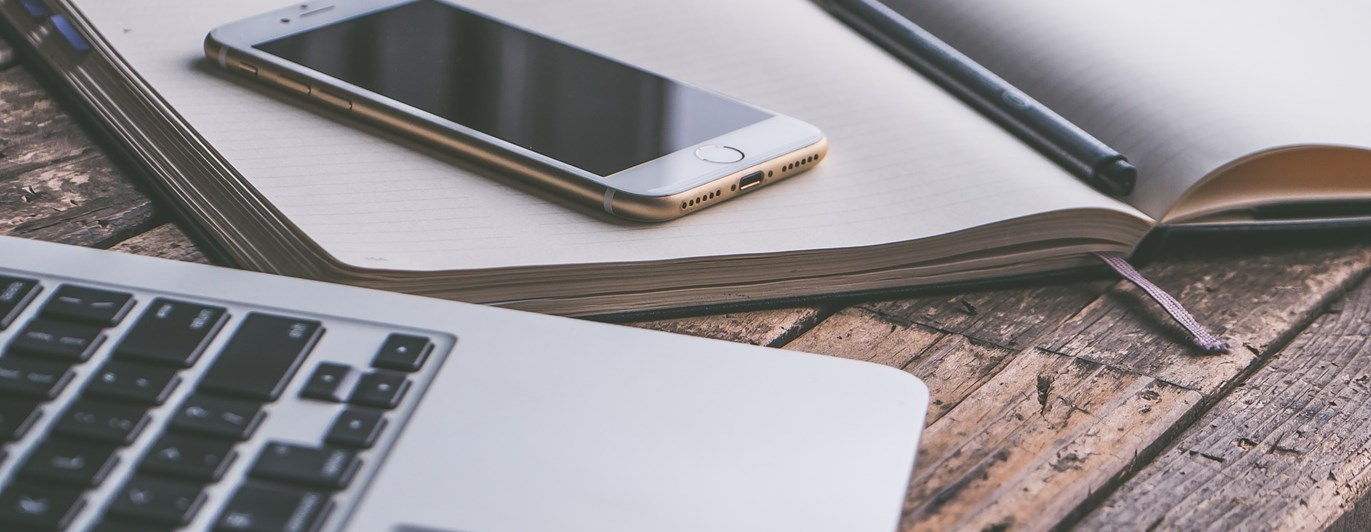 Bild som visar en bärbar dator, mobil och anteckningsblock på ett träbord.