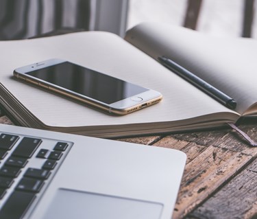 Bild som visar en bärbar dator, mobil och anteckningsblock på ett träbord.