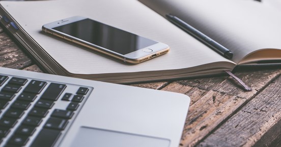 Bild som visar en bärbar dator, mobil och anteckningsblock på ett träbord.