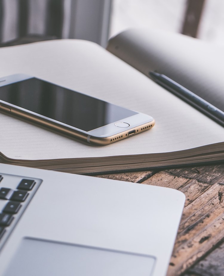 Bild som visar en bärbar dator, mobil och anteckningsblock på ett träbord.